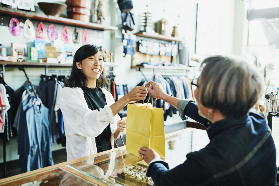 Shop owner handing bag to smiling client after shopping in clothing boutique