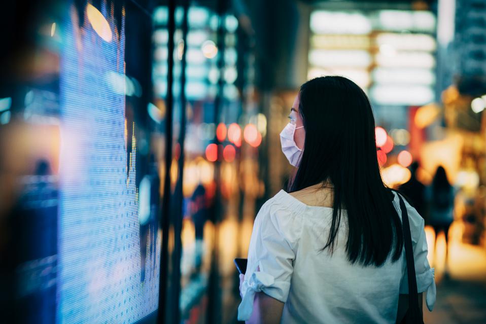 Economic and financial impact during the Covid-19 health crisis deepens. Businesswoman with protective face mask checking financial trading data on smartphone by the stock exchange market display screen board in downtown financial district showing stock m