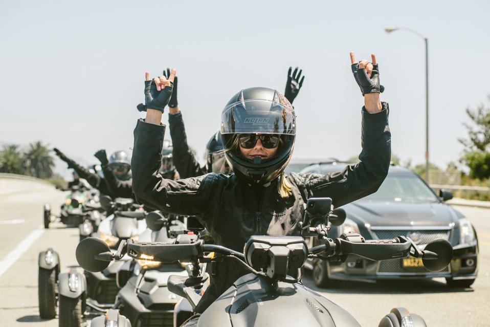 Five women pose on three-wheeled motorcycles on the side of a highway.
