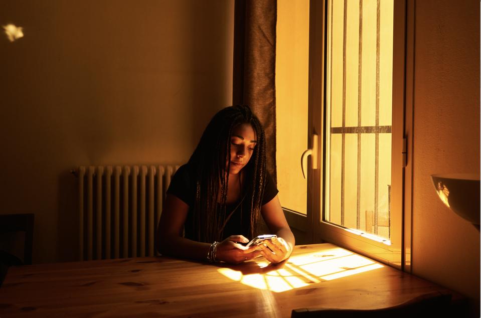 Aisha Coulibali co-founder of Mygrants sitting at a window with the sun shining at the table in front of her while she is looking at her phone