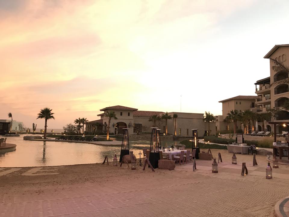 At sunset, a groomed sandy beach next to the lagoon pool, with lanterns lighting a path to a table set for dinner. In the background are buildings for Grand Solmar at Rancho San Lucas's main restaurant and guest suites.