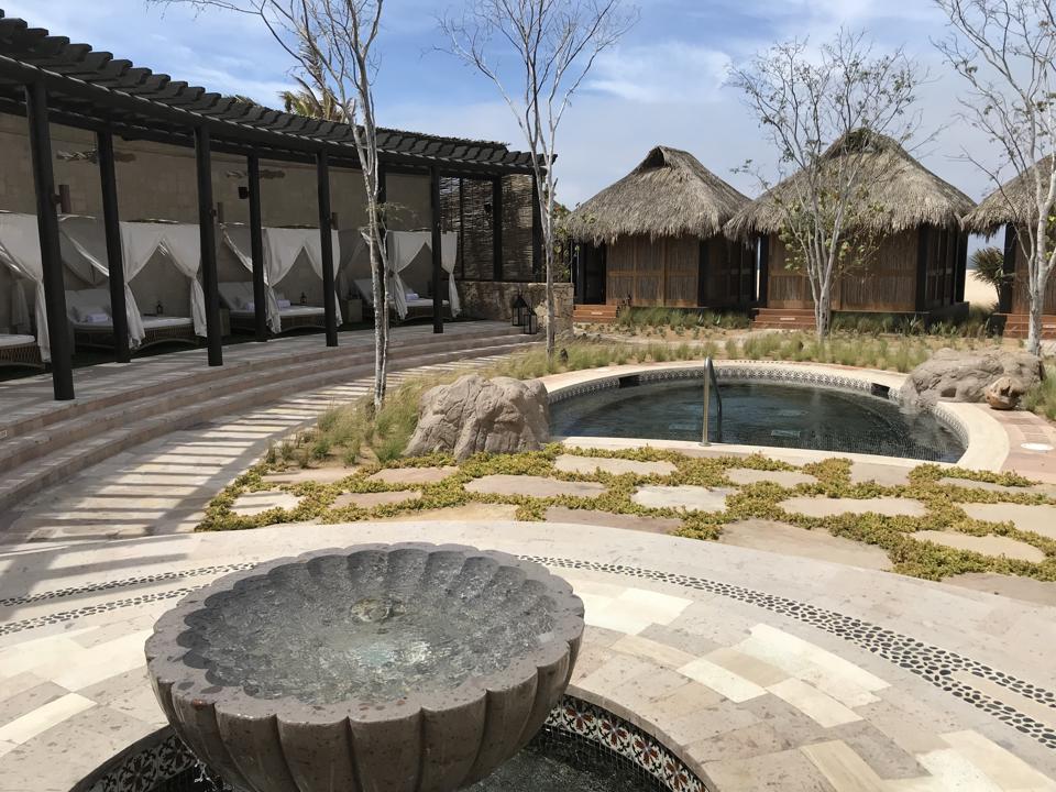 Three beachfront cabanas for spa treatments, to the left is a lounge area with curtained lounge beds, also in the photo are a small pool and a fountain