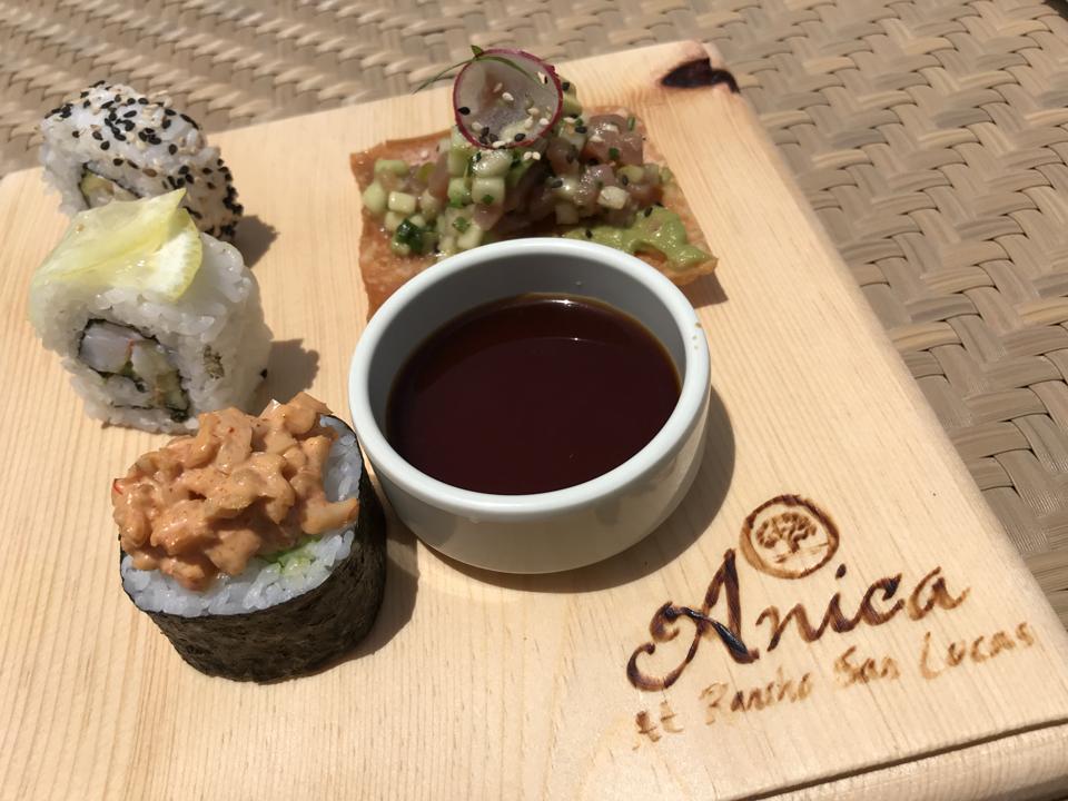 A wooden board with Anica's name burned into it, on top of which sits pieces of sushi, a ramekin of soya sauce, and a ceviche-topped square cracker