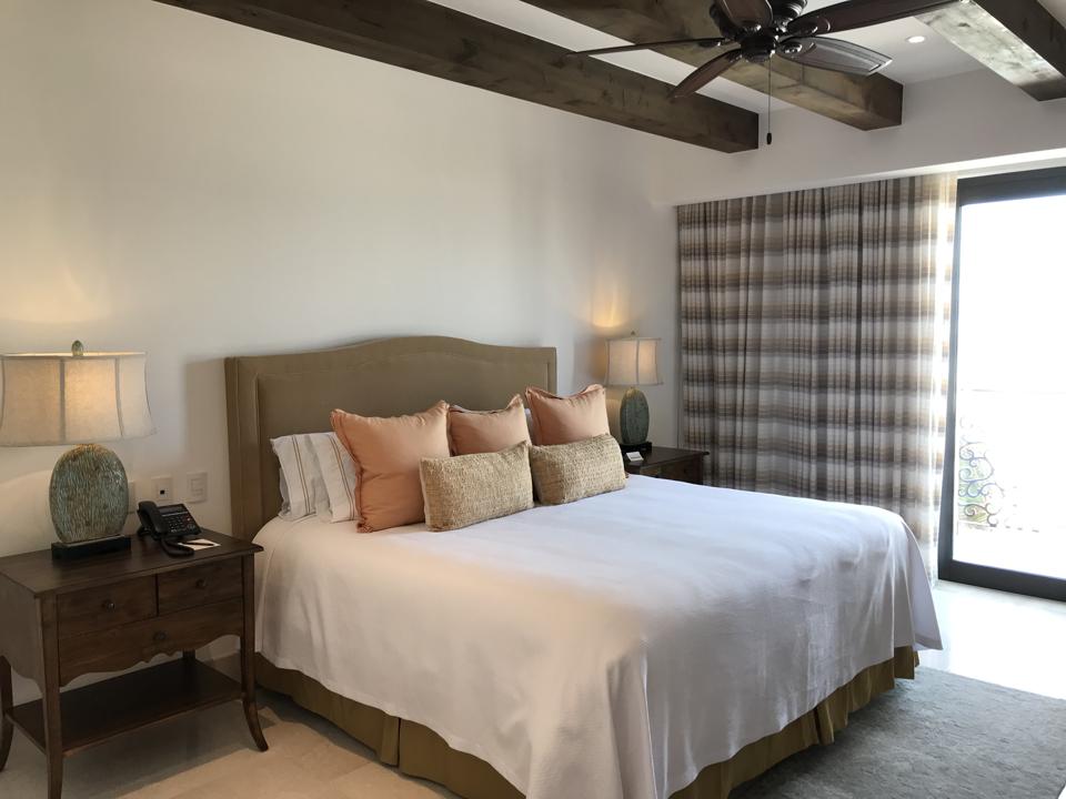 A king-sized bed with white cover and earth-toned throw pillows, with a bedside table, curtained doors go out to a balcony, and the ceiling has wooden beams and a fan