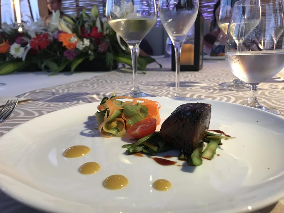 A plate with artfully-arranged beef and salad, on a white tablecloth with wine glasses and flowers.