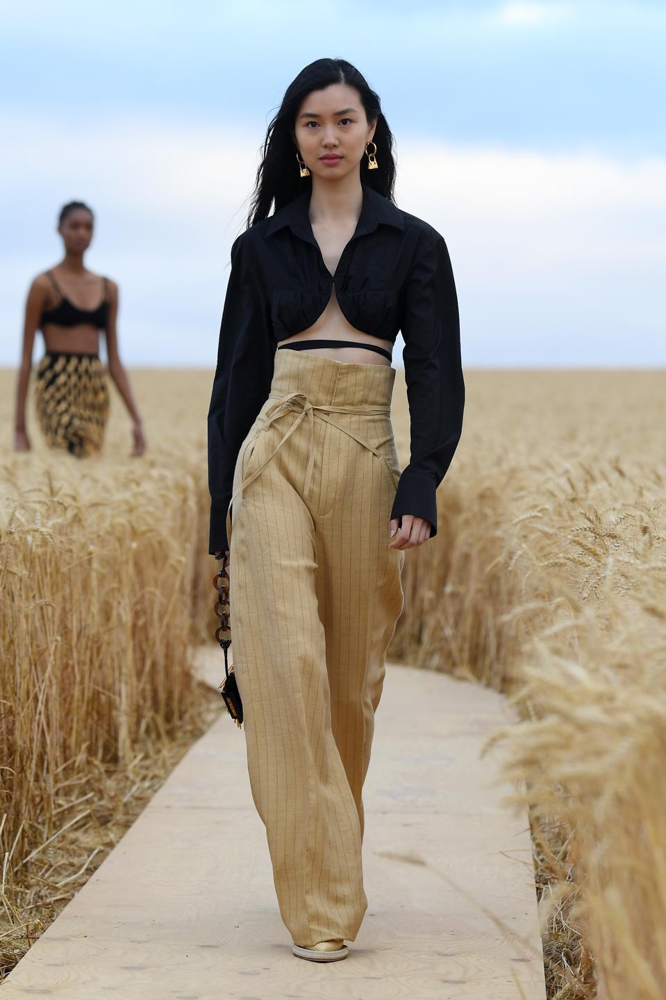 A model in a black cardigan and tan pants walks on a runway in a wheat field