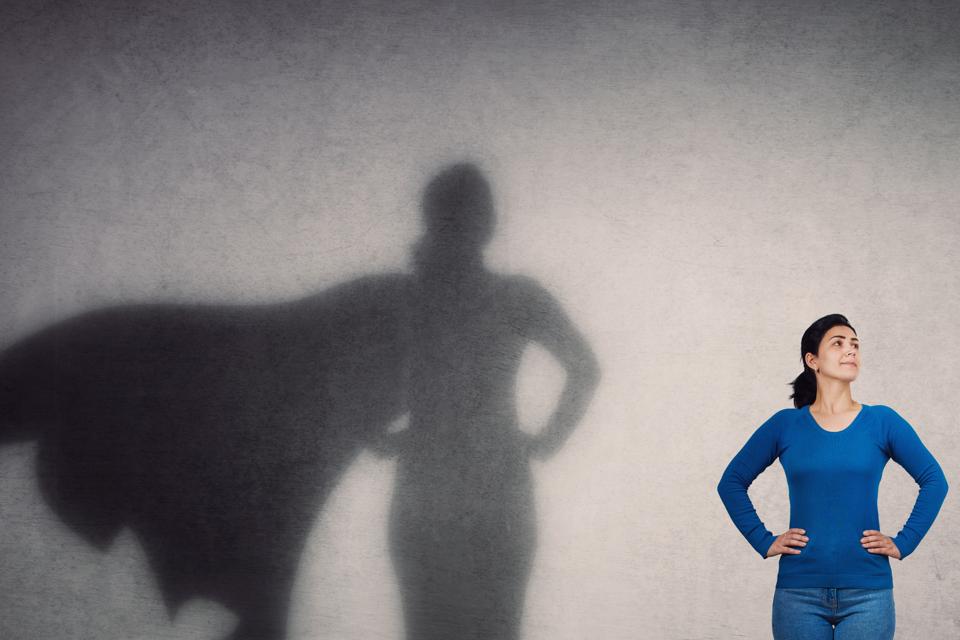 Brave woman keeps arms on hips, smiling confident, casting a superhero with cape shadow on the wall. Ambition and business success concept. Leadership hero power, motivation and inner strength symbol.