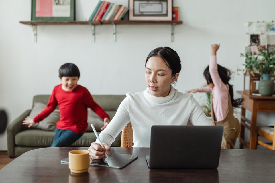 A woman works on a computer at home with her children playing in the background.