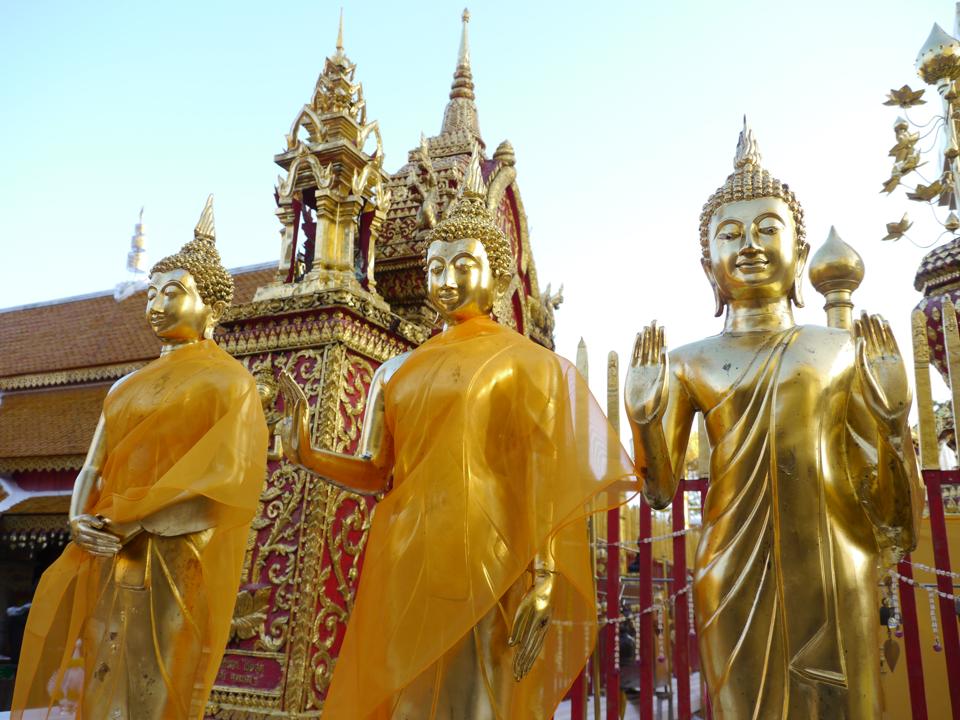 Three gold Buddha statues, two draped in orange and two with hands raised, in front of a gold and red temple