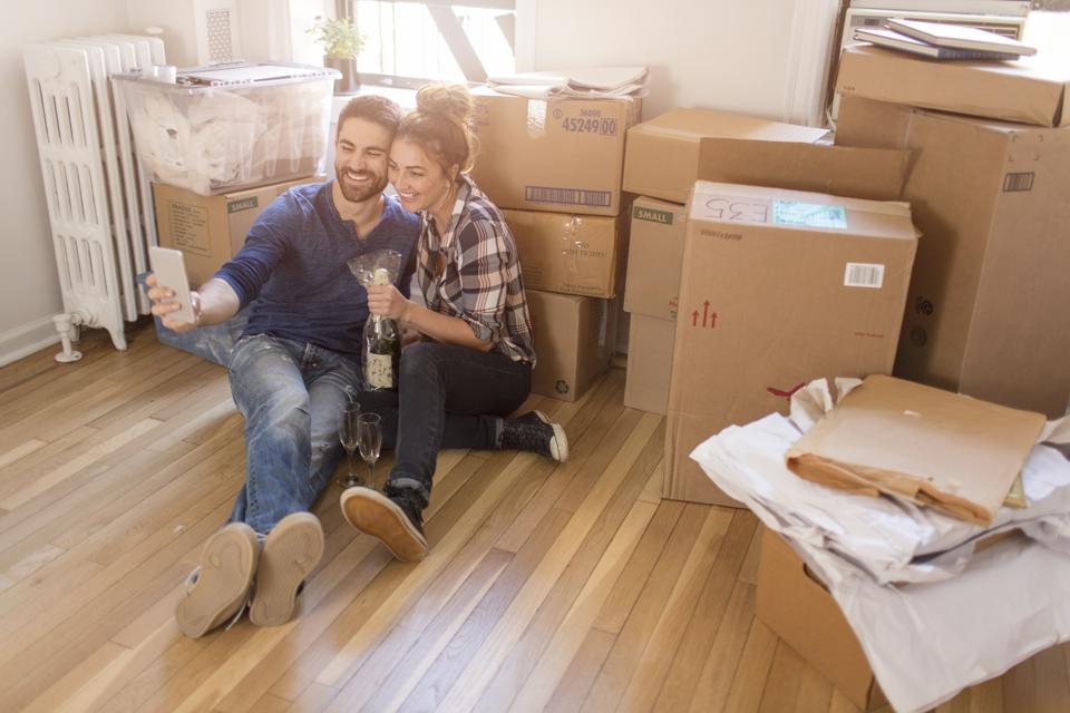 Moving house: Young couple sitting in room full of boxes, holding champagne bottle, taking self portrait with smartphone