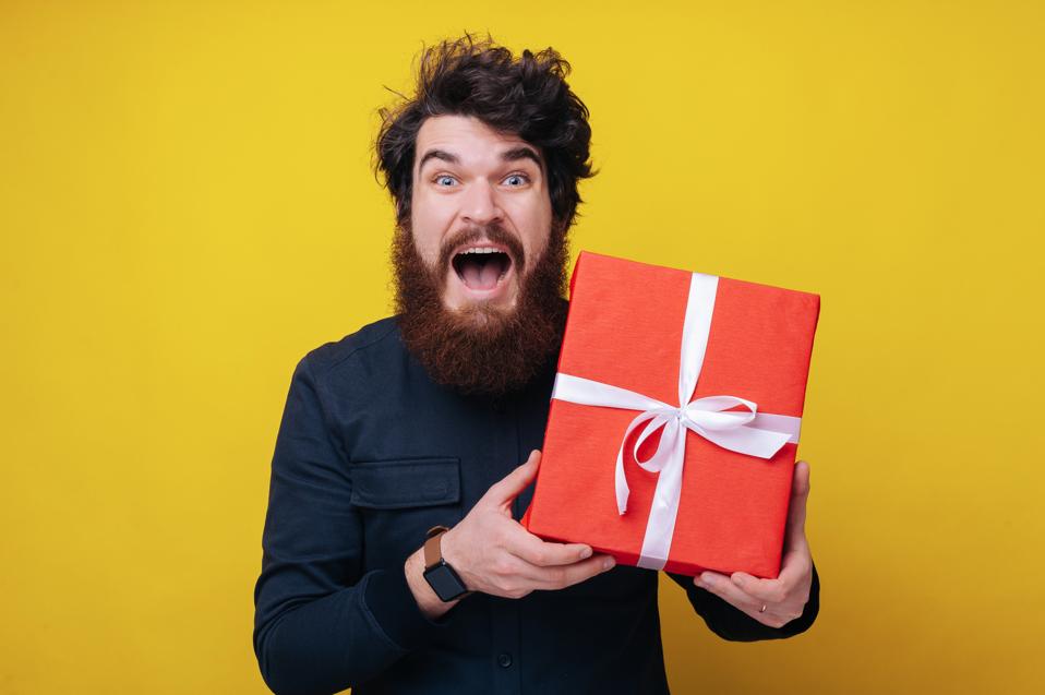 Handsome bearded man, looking excited at camera, holding a gift box, and standing over yellow background