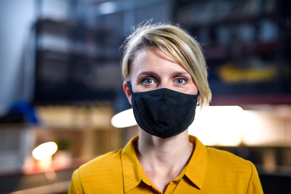 Portrait of woman with face mask standing indoors in office, looking at camera.