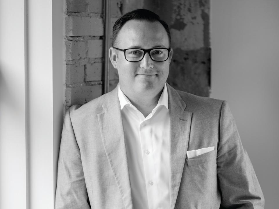 A man wearing black rimmed glasses, a white shirt and grey suit coat.