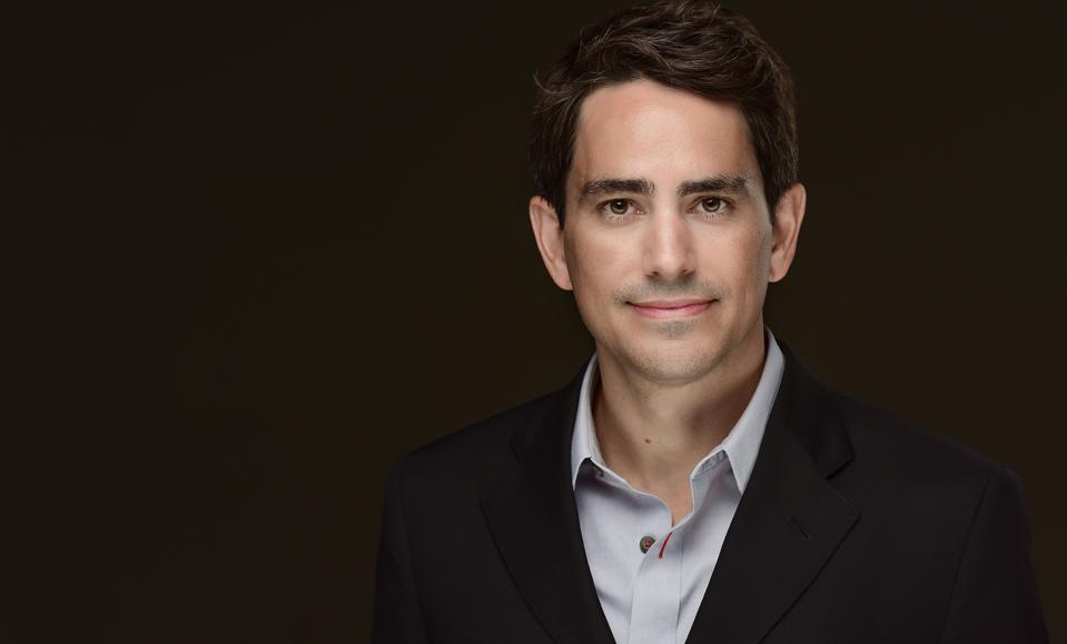 Brown haired business man wearing a suit coat, smiling with lips pursed.