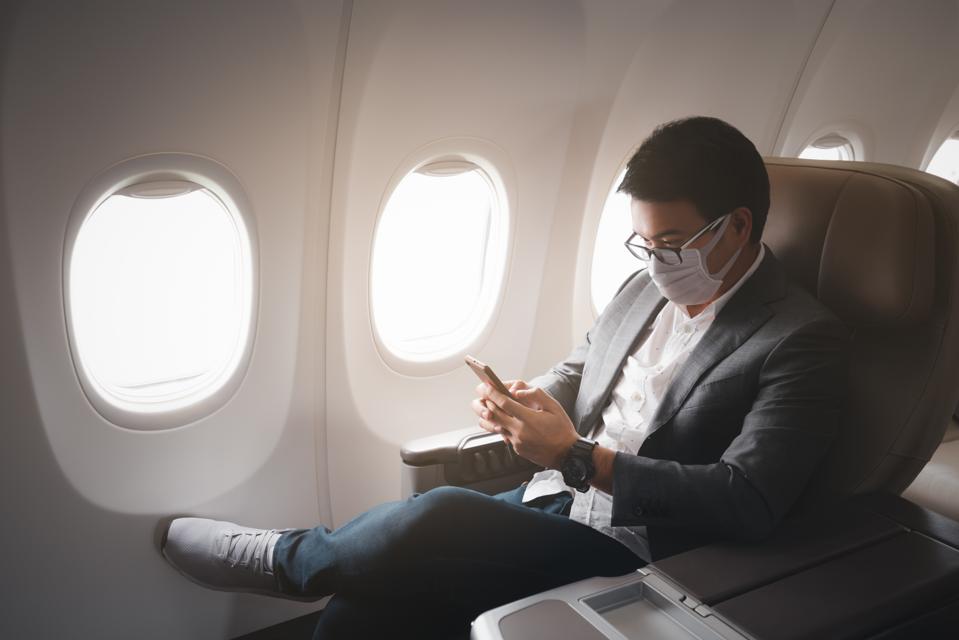 Young asian businessman wearing protective face mask with suit sitting in business first class seat using smartphone due to Coronavirus or COVID-19 outbreak situation in all of landmass in the world