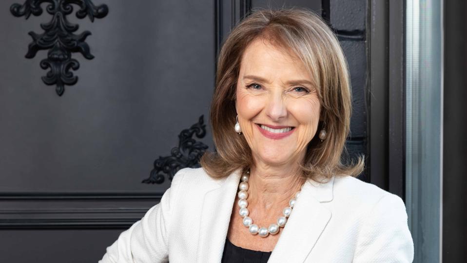 Business woman wearing white business jacket smiling at camera leaning against window.