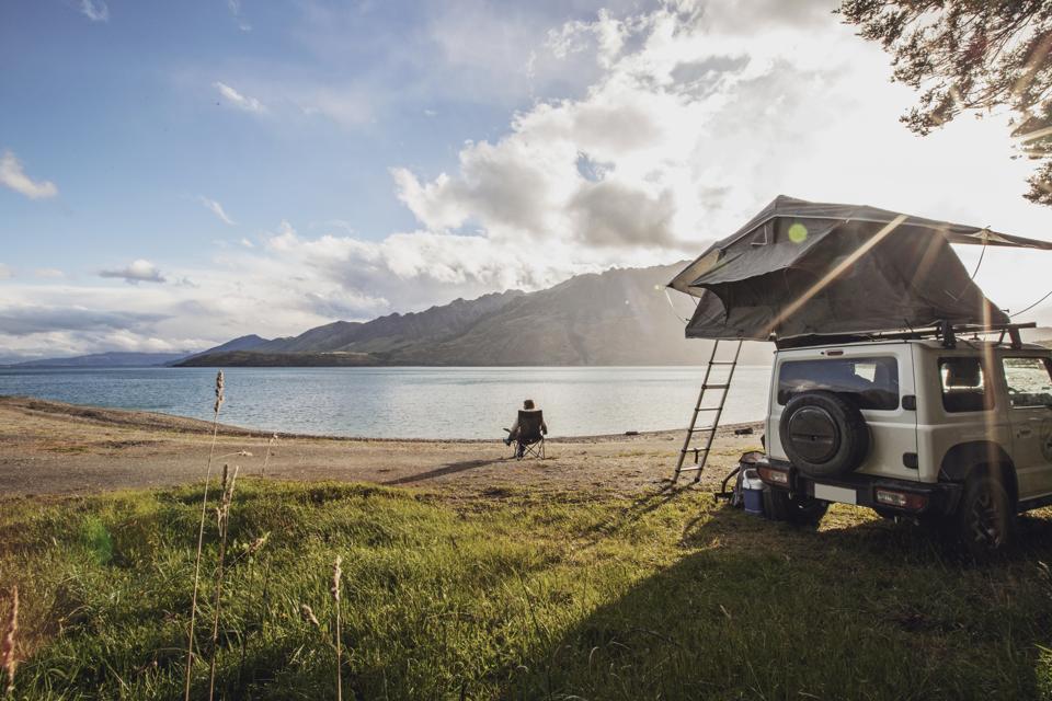 A woman sits along a lake next to a SUV with a tent in New Zealand