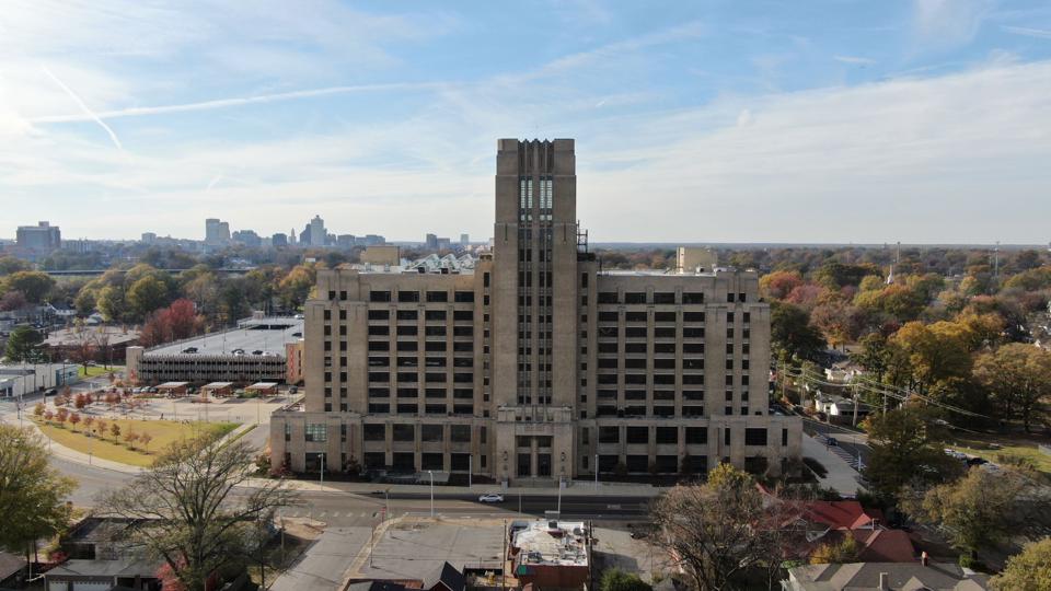 A large, beige warehouse building with a bell tower protrudes from an urban forest.