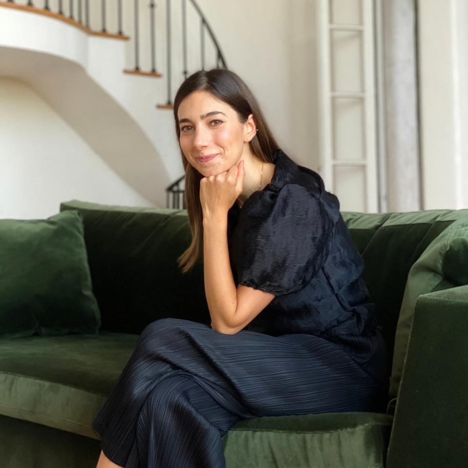 A white woman with brown hair sits on a green couch in a black dress.