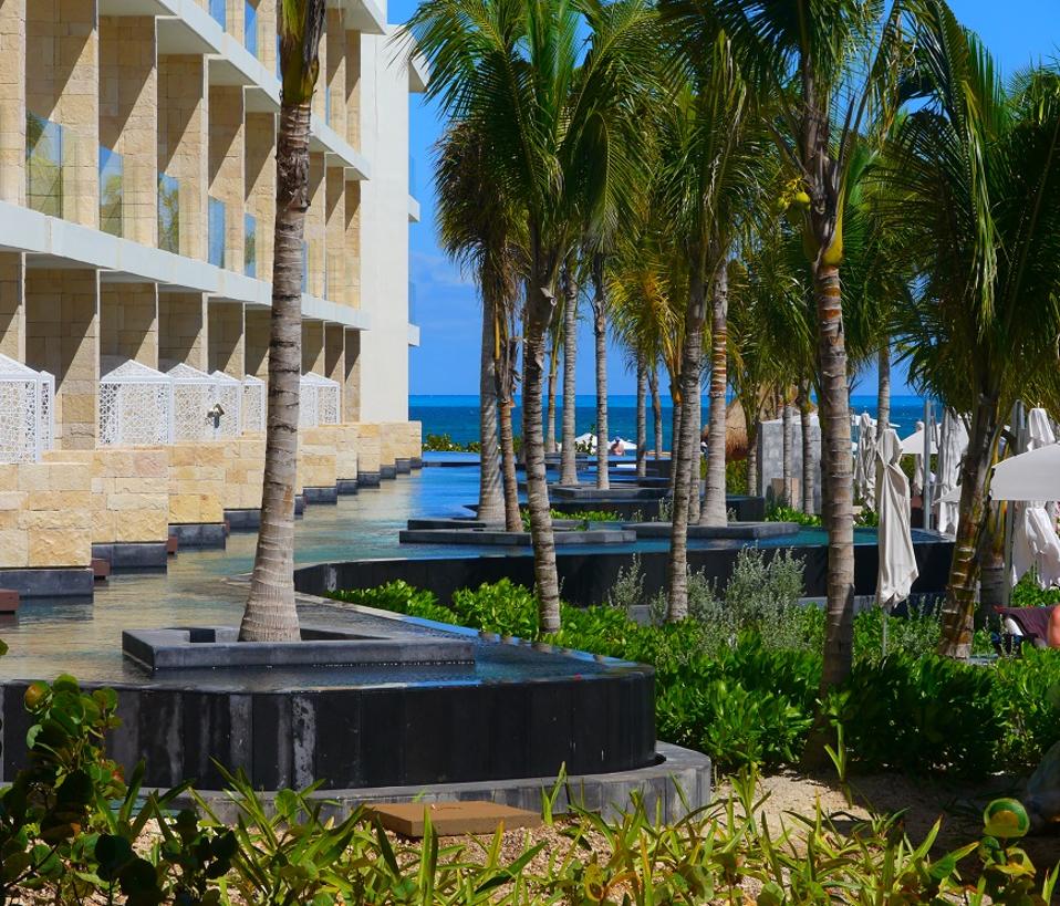 Palm trees line a swim-up pool in front of several hotel suites; the ocean is in the background.   