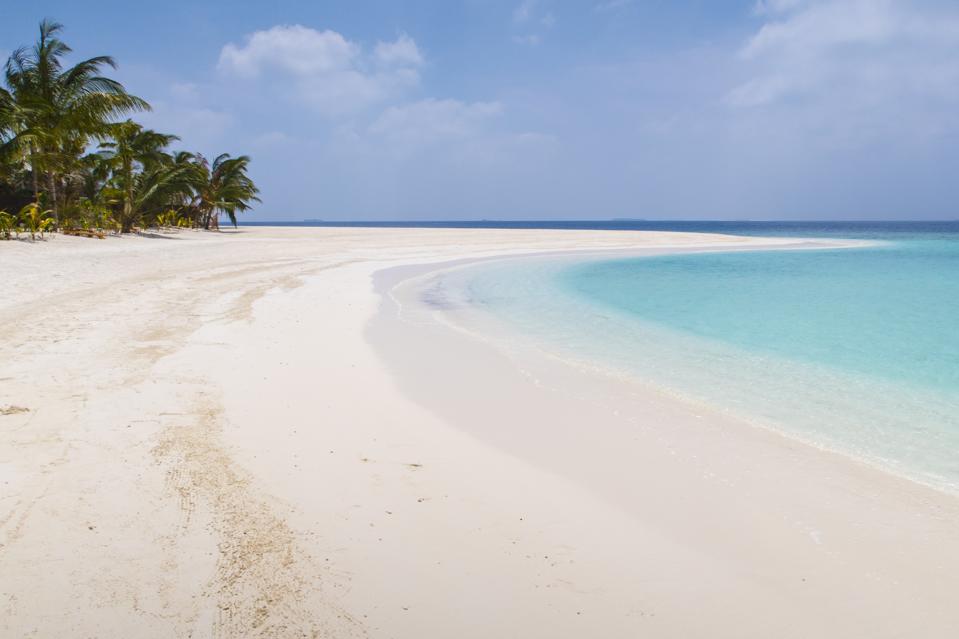 A wide, white sand beach with palm trees curves next to clear blue water.