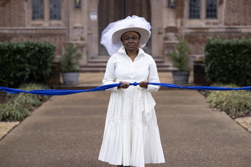 A woman in a white dress and hat stands outside of a large building holding a blue ribbon.