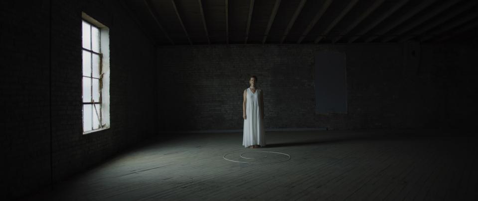 A woman in a white dress stands in a dark room, lit by a single window.