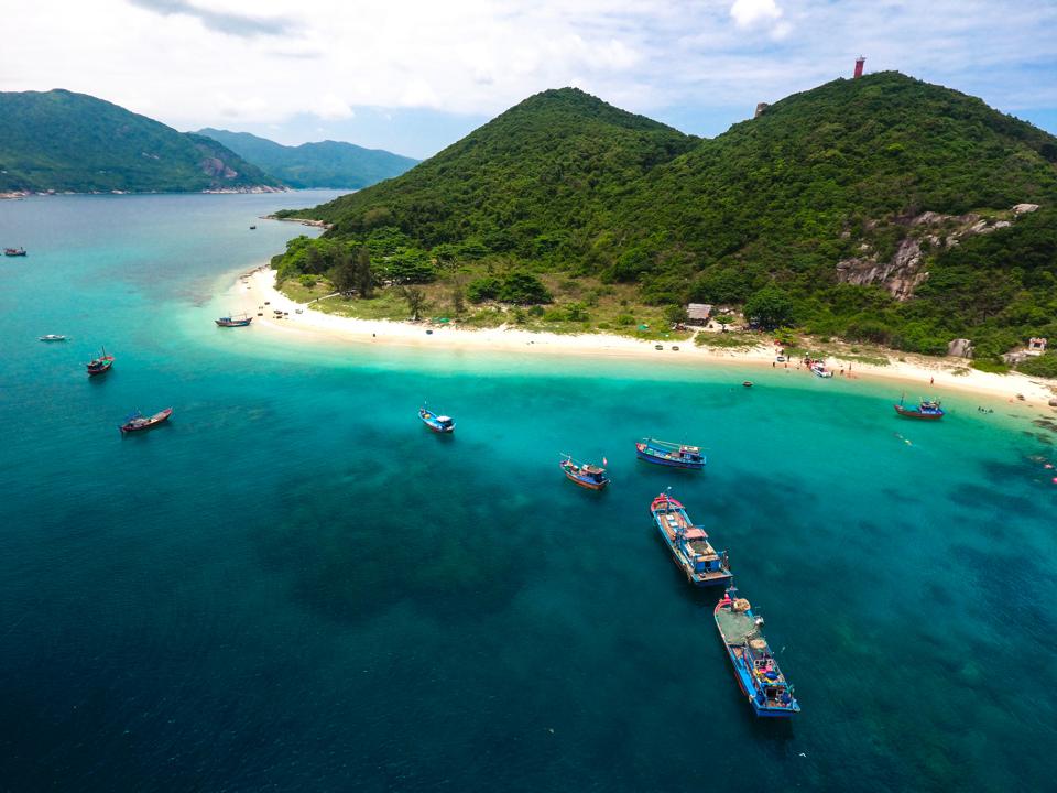 A hilly island surrounded by colorful barges and turquoise water. 