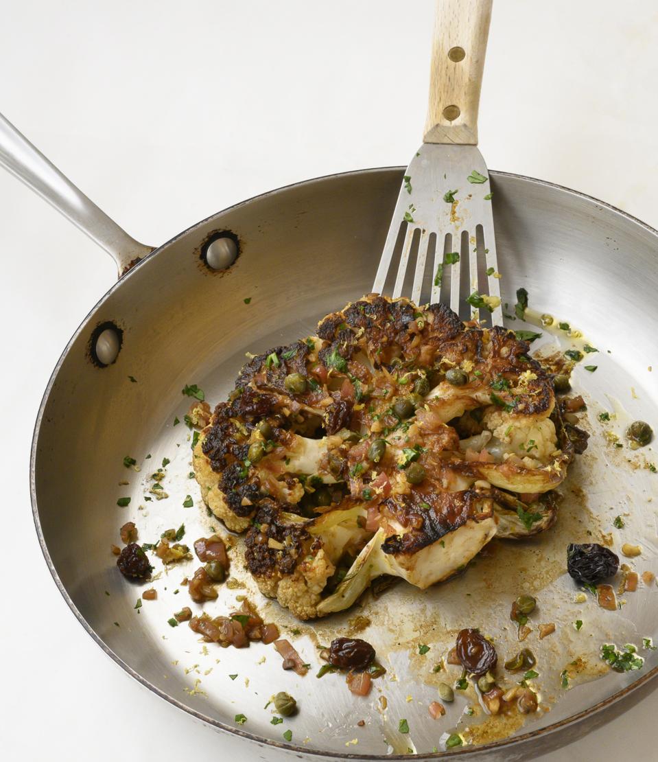 A sautéed cauliflower steak with a lemon, anchovy, caper sauce is photographed in a stainless-steel sauce pan with a stainless spatula lifting it up. The photograph is from ″Flavorbomb″ cookbook by Bob Blumer.