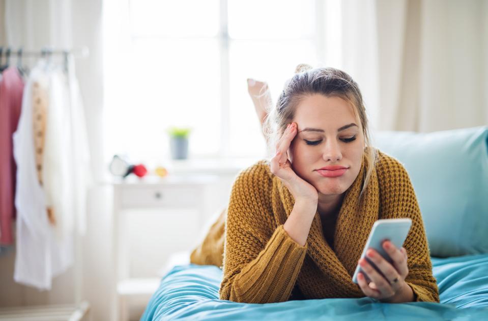 Bored young woman with smartphone lying on bed indoors at home.