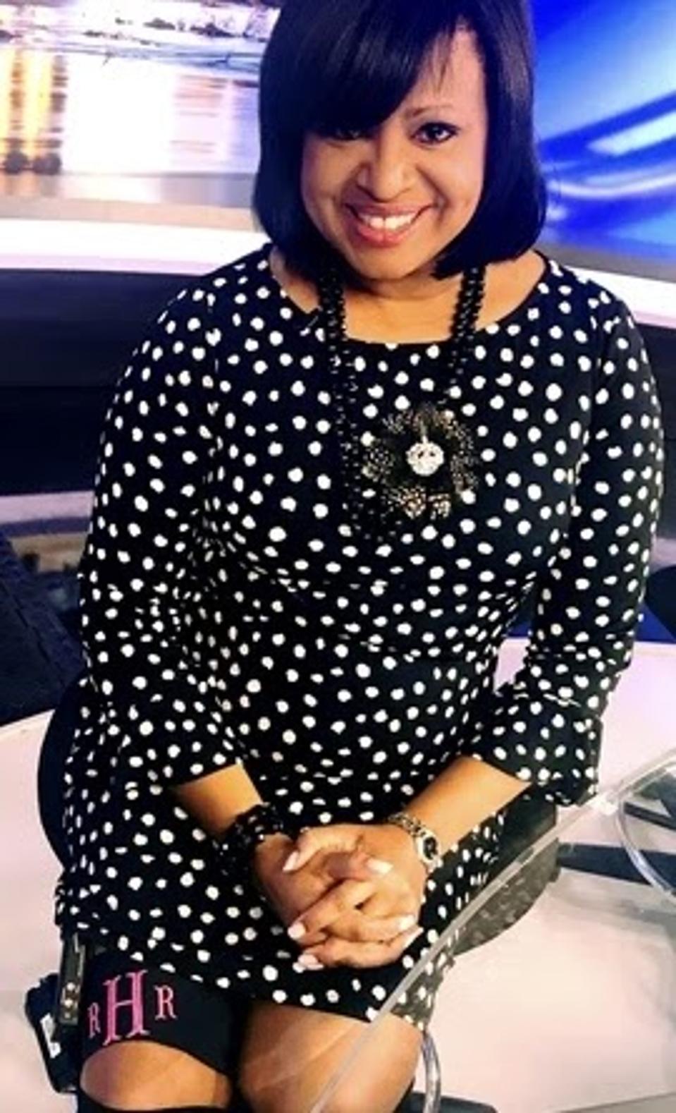 A Black woman in a black and white polka dot dress smiles on a television news set.