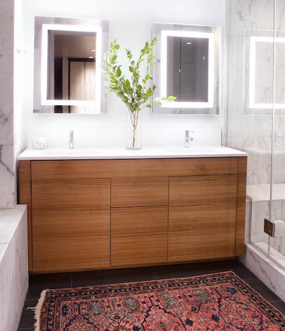 a marble bathroom with a red Persian rug, lit mirrors and wood vanity