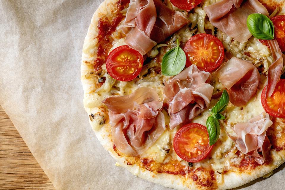 Fresh baking homemade pizza napolitana with prosciutto ham, cheese, tomatoes, basil on baking paper over wooden table background. Home baking or delivered fast food. Flat lay, space.