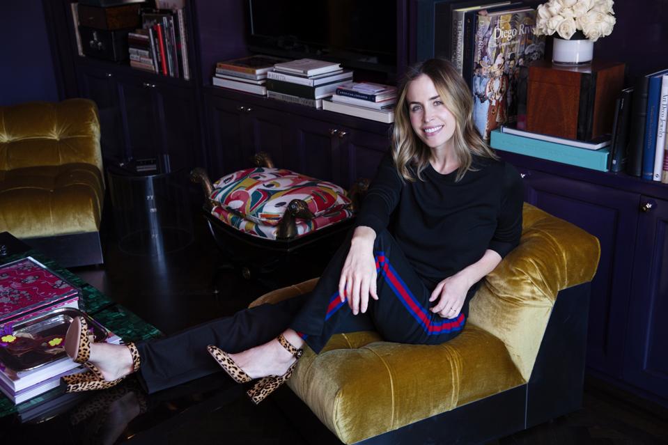 Designer Brett Heyman in a black outfit with leopard print shoes on a velvet chair.