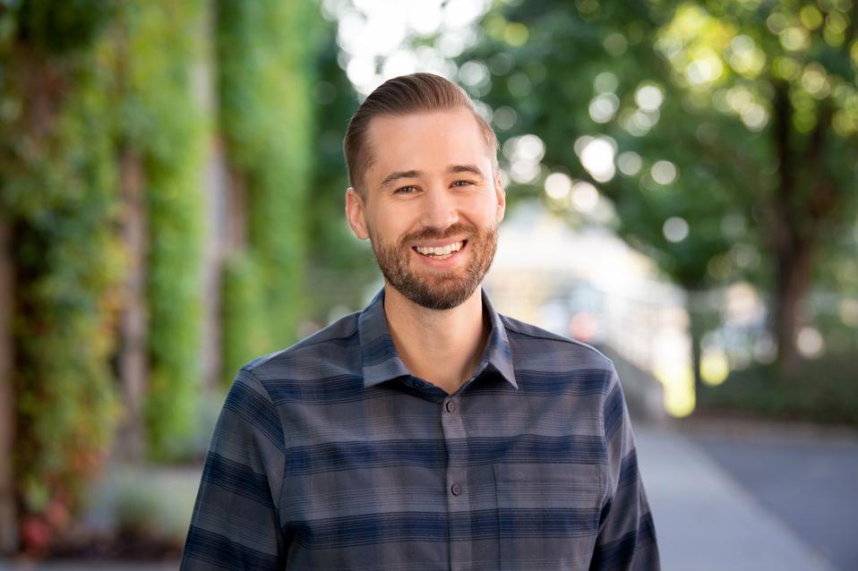 A young white man with beard wearing a plaid shirt smiling into camera