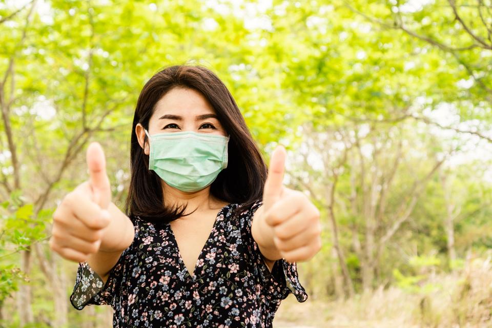 Asian woman wearing face mask walking outdoors to prevent virus covid-19 with thumbs up.