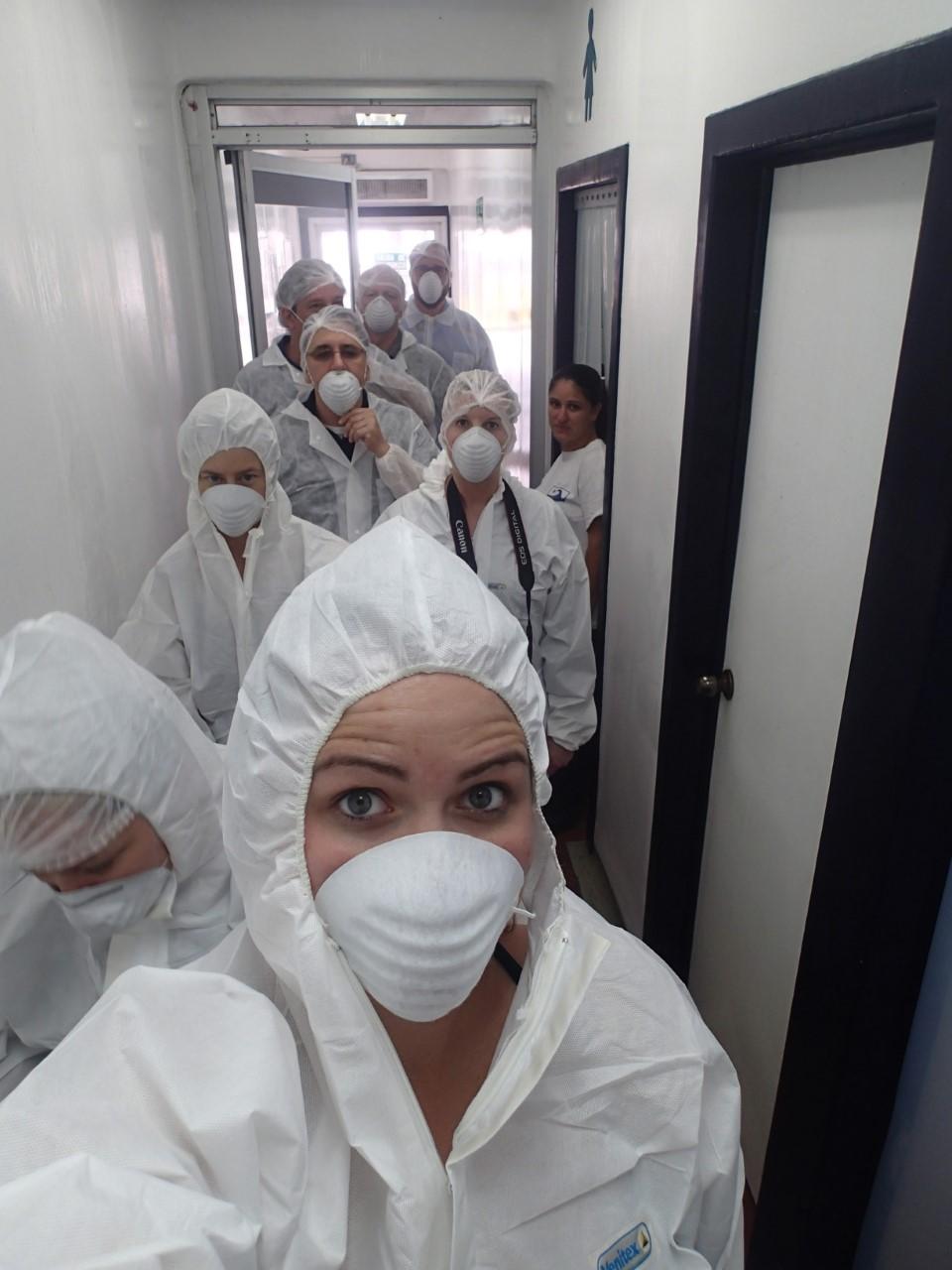 Woman in PPE and lab coat takes a selfie looking into the camera with 8 similarly-dressed individuals behind her in a hallway.