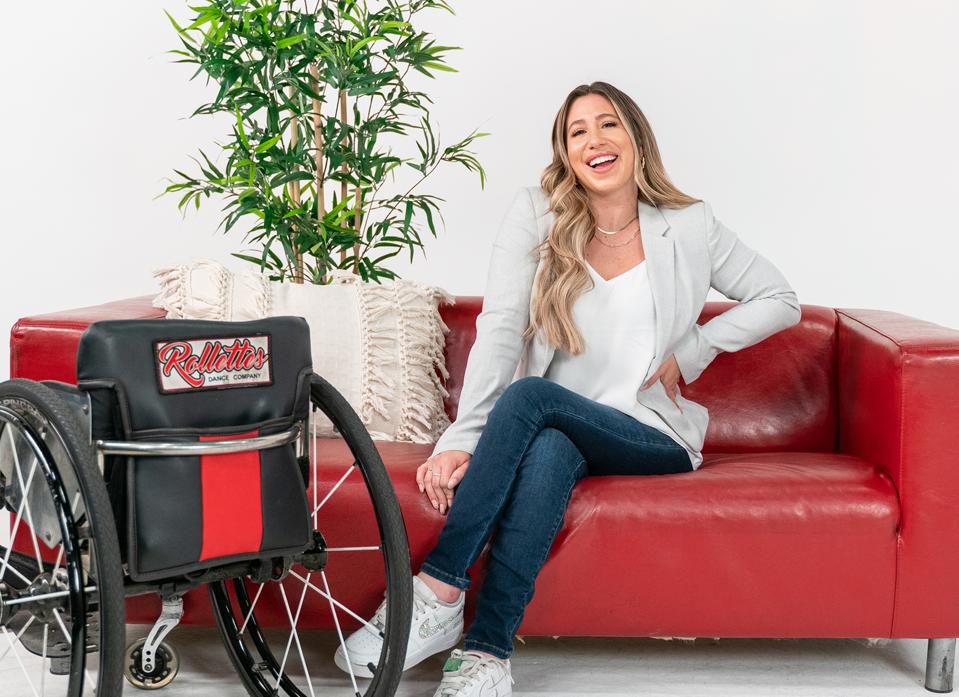 Chelsie Hill sits on a red couch smiling next to a Rollettes wheelchair.