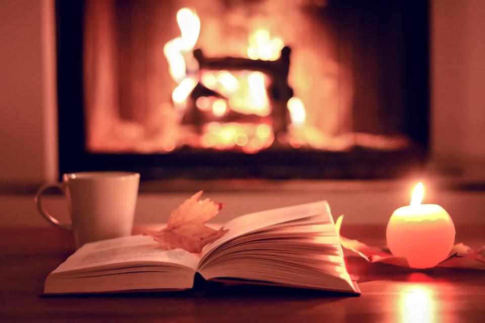 Cozy evening with fireplace at home. Close up photo of open book with leaf, cup of tea and candle with fire at background.