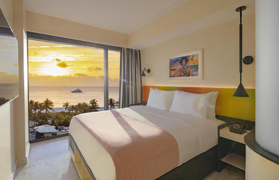 A sunny hotel bedroom with a floor-to-ceiling window offering unobstructed views of the nearby ocean.
