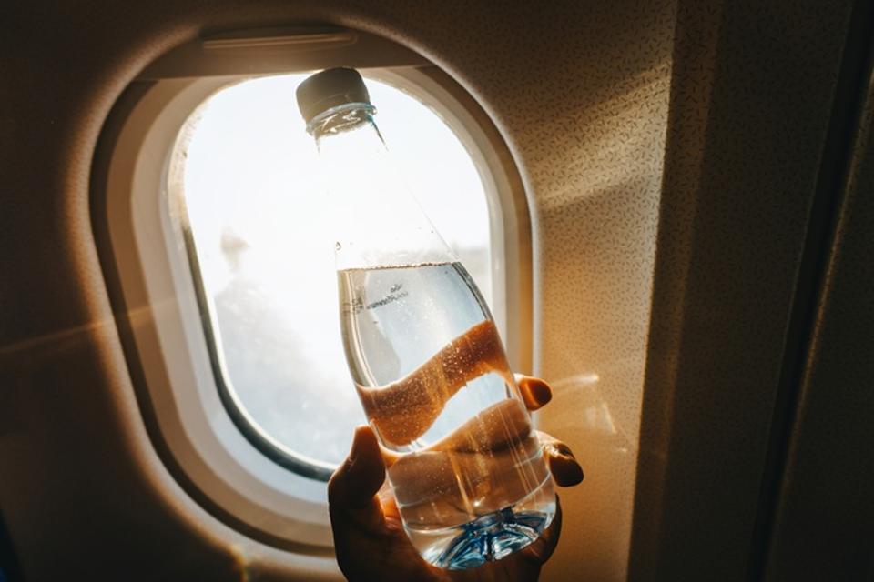 Cropped hand of woman holding a bottle of water against airplane window in front of golden sunbeam while travelling.