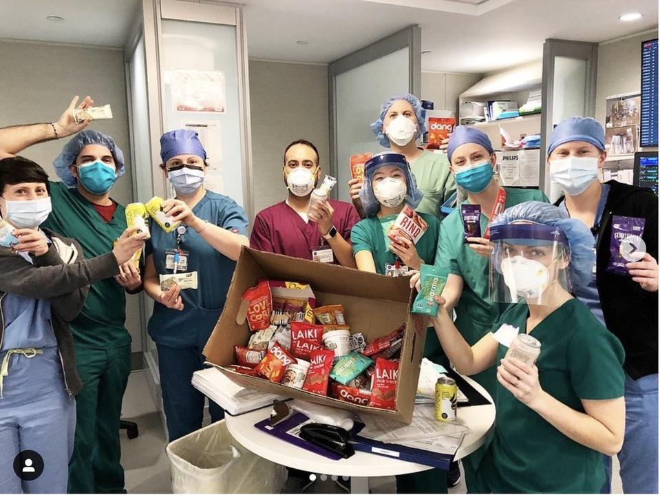 A group of 10 healthcare workers in scrubs and caps hold up snacks from a large box