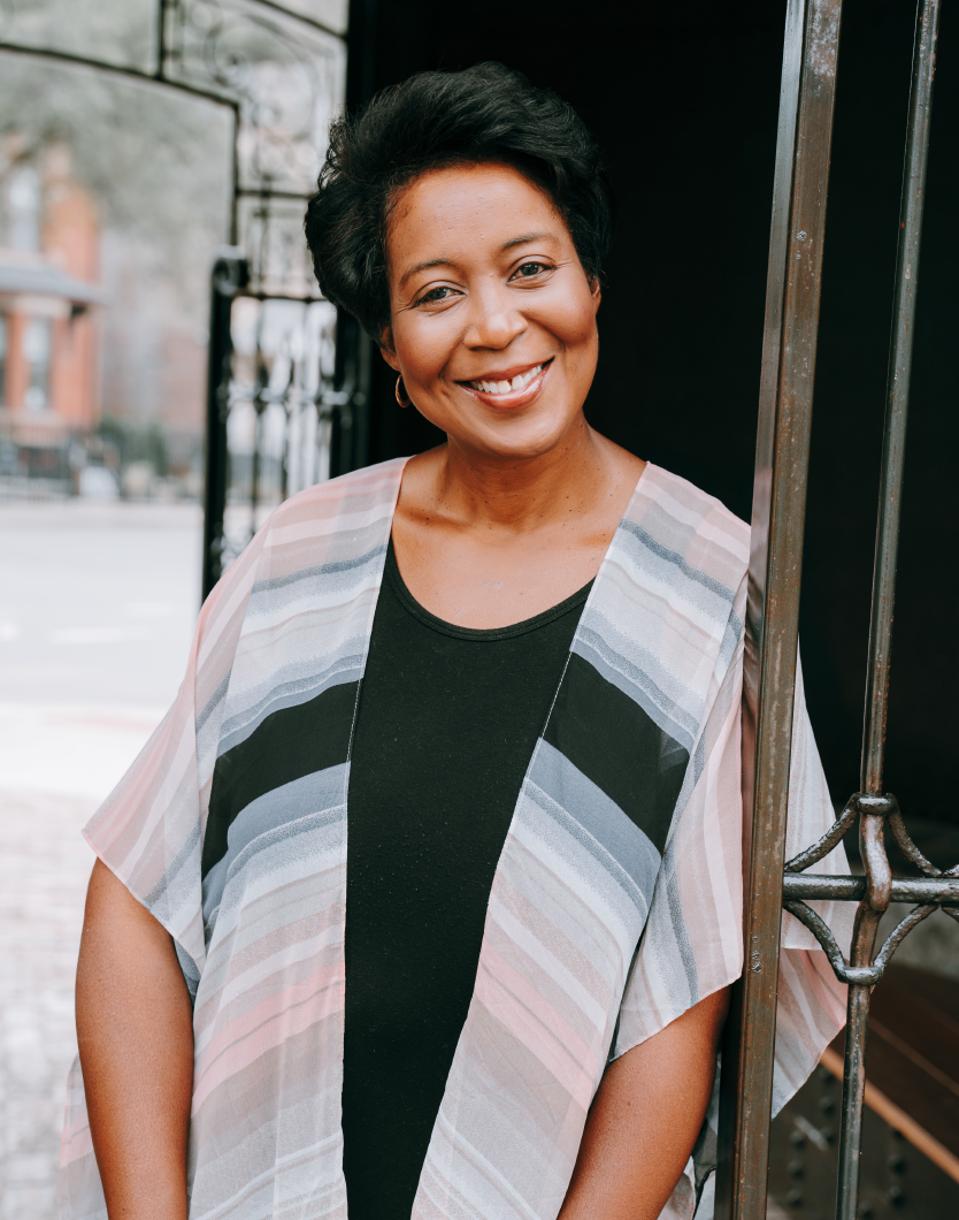 A dark-haired woman, Devita Davison, stands smiling, wearing a black top and striped shawl.
