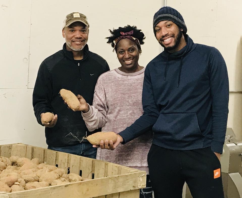 Two Black men and a Black woman stand holding sweet potatoes.