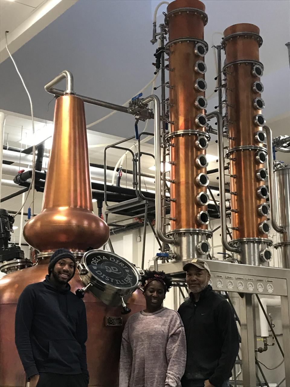 Three people standing in front of large columns of distilling equipment.