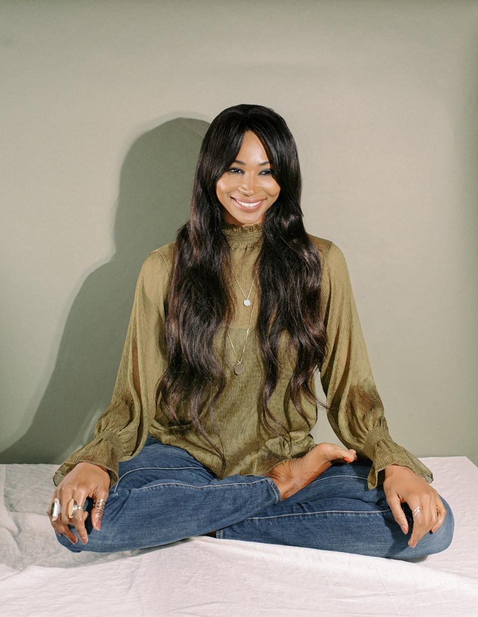 A Black woman sits with her legs crossed. She has long brown hair.
