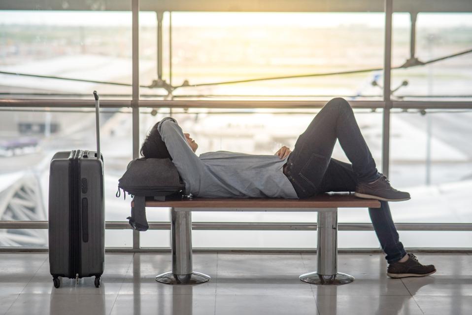 Young Asian man with suitcase luggage and backpack lying on bench in airport terminal