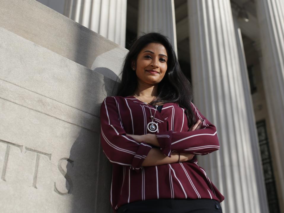 Shriya Srinivasan shown standing smiling leaning against an MIT building with columns.