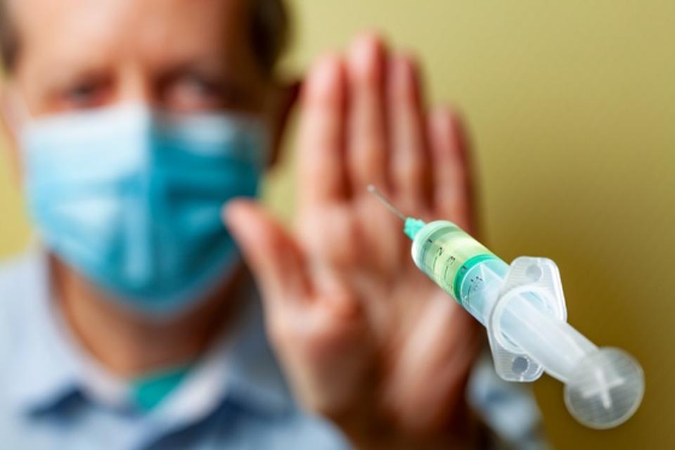 Holding up hand to stop a COVID-19 vaccination syringe from approaching, an anti vaxxer person stands in the background