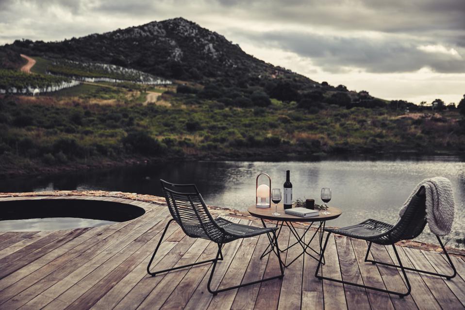 Two black chairs on a wood deck with wine glasses, a wine bottle facing the water